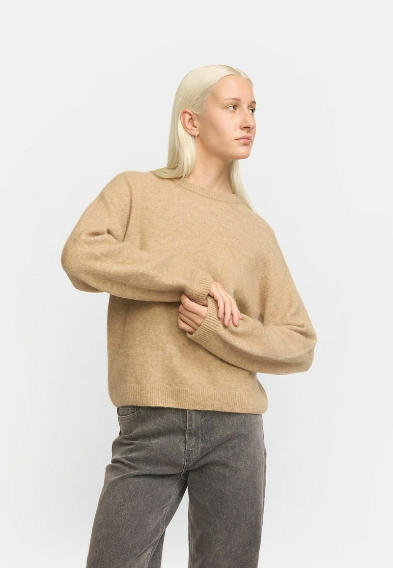 Beige gestrickter Pullover mit Rundhalsausschnitt, gerippten Bündchen und Saum sowie einer weichen Textur, kombiniert mit grauen hoch geschnittenen Jeans.