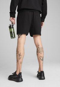 Șorturi atletice negre cu un tiv elastic, ținute cu o mână. În cealaltă mână este o sticlă de apă verde, cu tatuaje vizibile pe picioare.