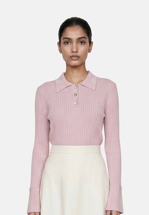 Donna con capelli scuri che indossa una polo rosa a coste con maniche lunghe e bottoni, in piedi davanti a uno sfondo chiaro e uniforme.