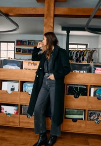 Lang svart frakk med store frontknapper, kombinert med en mørk denimskjorte og grå vide jeans. Står ved siden av en treoppbevaring for plater.