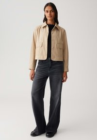 Donna in piedi con una giacca beige con bottoni sopra una maglietta nera, jeans a gamba larga neri sbiaditi e scarpe nere, su uno sfondo semplice.
