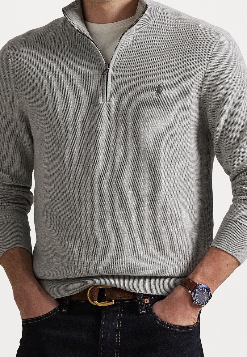 Pull-over gris texturé avec un col à fermeture éclair et des poignets côtelés. Porté sur une chemise claire, associé à un jean foncé et une ceinture marron.