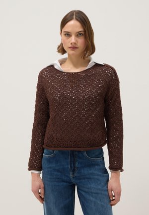 Donna che indossa un maglione marrone a maniche lunghe lavorato a uncinetto sopra una camicia bianca con colletto e jeans blu, in piedi contro uno sfondo semplice.