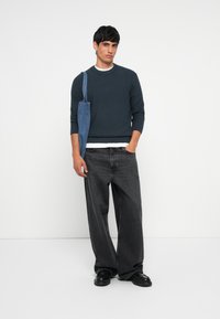 Dark green ribbed sweater, white shirt underneath, wide black jeans, and black shoes. A blue denim bag hangs from his shoulder.
