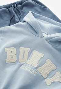 A világos kék kapucnis pulóver fehér hímzett "BUNNY ACADEMY" felirattal rendelkezik. Kék melegítőnadrág látható alatta, gumis derékkal.