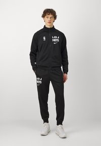 Tuta sportiva nera realizzata in un tessuto morbido. Presenta il logo dei NBA Nets e il logo Nike in bianco. Giacca con zip, polsini aderenti e vita elastica.
