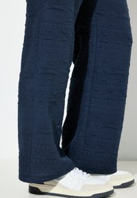 Pantaloni texture blu navy con un motivo geometrico, abbinati a sneakers bianche. Il tessuto appare morbido con un design leggermente in rilievo.