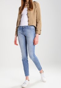 Kobieta w beżowej, zapinanej na guziki kurtce, białym topie na ramiączkach, jasnoniebieskich, dopasowanych dżinsach oraz białych wsuwanych trampkach, stojąca na jednolitym tle.