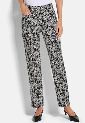 Frau trägt schwarze Hose mit weißem Blumenmuster, ein weißes Oberteil und schwarze spitze Pumps, steht mit einer Hand in der Tasche.