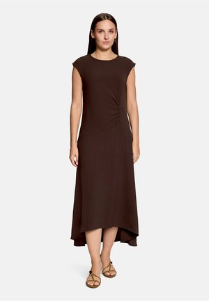 Femme aux cheveux bruns raides porte une robe sans manches marron foncé avec un ourlet asymétrique et des fronces sur le devant, associée à des sandales couleur beige.