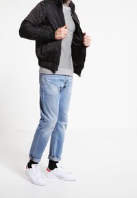 Svart quiltad jacka, grårandig skjorta, ljusblå jeans och vita sneakers med röda accenter. Avslappnad passform med uppvikt jeanstyg.