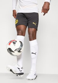 Pantaloni neri con logo giallo del Borussia Dortmund e branding Puma, abbinati a calze bianche. Un pallone da calcio con motivo bianco e nero è tenuto all'altezza del ginocchio.