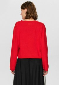 Maglione rosso corto con maniche lunghe, texture morbida e scollatura ampia, abbinato a una gonna nera con sovrapposizione trasparente.
