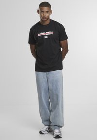 Schwarzes T-Shirt mit der Aufschrift "MÜNCHEN" in roten Blockbuchstaben, kombiniert mit hellblauen lässigen Jeans und grauen Turnschuhen. Einfaches Design, aus Baumwolle.