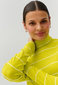 Svetlo rumena rebrasta turtleneck s belimi črtami. Tkanina prikazuje teksturiran vzorec. Model nežno počiva z roko na vratu.