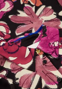 Bloemige blouse van zwarte stof, met grote roze en crèmekleurige bloemen, een blauwe binnenkraag en een gouden accent in de vorm van een bloem.