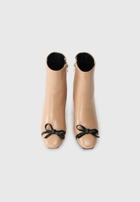 Μπεζ μποτάκια μέχρι τον αστράγαλο με γυαλιστερή επιφάνεια, που διαθέτουν μια μαύρη κορδέλα στην άκρη και φερμουάρ στο πλάι για κλείσιμο. Σχεδίαση τετράγωνης μύτης.