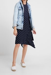Námořnické šaty s puntíky a asymetrickým spodním lemem, doplněné světle modrou denimovou bundou. Modelka má na sobě bílé espadrilové balerínky.