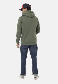 Haut à capuche vert olive avec une grande capuche, des poignets et un ourlet côtelés, associé à un jean bleu foncé et des chaussures à motif damier.