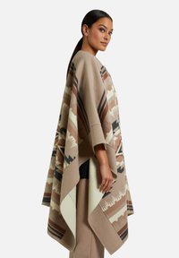 Beiger Poncho mit geometrischem Muster in Braun und Schwarz. Weiche Textur, lockere Passform, asymmetrischer Saum und breite Ärmel.