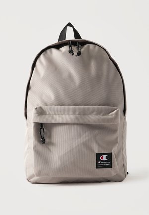 Sac à dos en tissu gris clair avec poche frontale zippée et écusson noir du logo Champion en bas à droite.