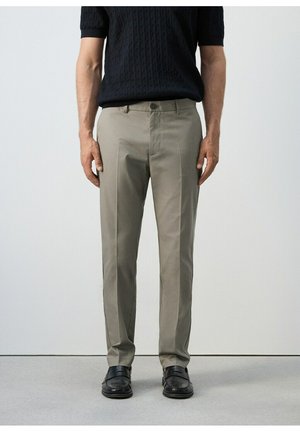 Uomo che indossa pantaloni beige, cintura nera, mocassini neri e un maglione nero a maniche corte con trama, in piedi contro uno sfondo semplice.