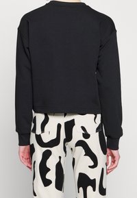 Personne portant un haut noir uni à manches longues et un pantalon blanc taille haute avec des motifs abstraits noirs irréguliers, vue de dos.