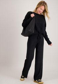Zwart gebreide trui en bijpassende brede broek met tunnelkoord in de taille, gestyled met een zwarte boodschappentas en zwart/geel sneakers.