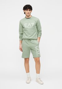 Groene katoenen sweater en korte broek set met witte geprinte logo's. De sweater heeft een ronde hals; de korte broek heeft een elastische tailleband en zakken.