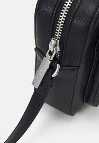 Borsa a tracolla in pelle nera con zipper in argento, strap regolabile e targhetta metallica. Superficie testurizzata con cuciture e dettagli in evidenza.
