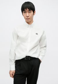 Biała koszula zapinana na guziki z czarnym logo w formie rysunkowej twarzy na piersi, zestawiona z czarnymi, dopasowanymi spodniami oraz czarnym paskiem.