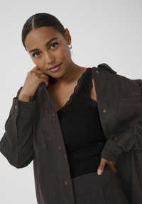 Mørkebrun ruskindsjakke med knaplukning, oversized pasform og skulderepauleter, Parret med en sort tanktop med blonder.