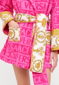 Ružičasti kupkaći ogrtač od frotira s odvažnim uzorkom logotipa "VERSACE". Bijeli satenasti ovratnik i remen ukrašeni zlatnim baroknim motivima.