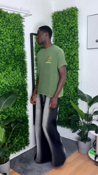 Camiseta de algodón verde con un logo de mariposa, combinada con pantalones de pierna ancha en negro y gris, en un entorno de vegetación artificial y plantas de interior.