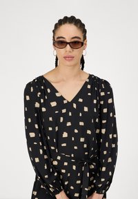 Μαύρο φόρεμα με μπεζ τετράγωνα σχέδια, V λαιμόκοψη, μακριά μανίκια με φούσκωμα στους ώμους και ζώνη στη μέση. Συνοδεύεται από σκούρα γυαλιά ηλίου.