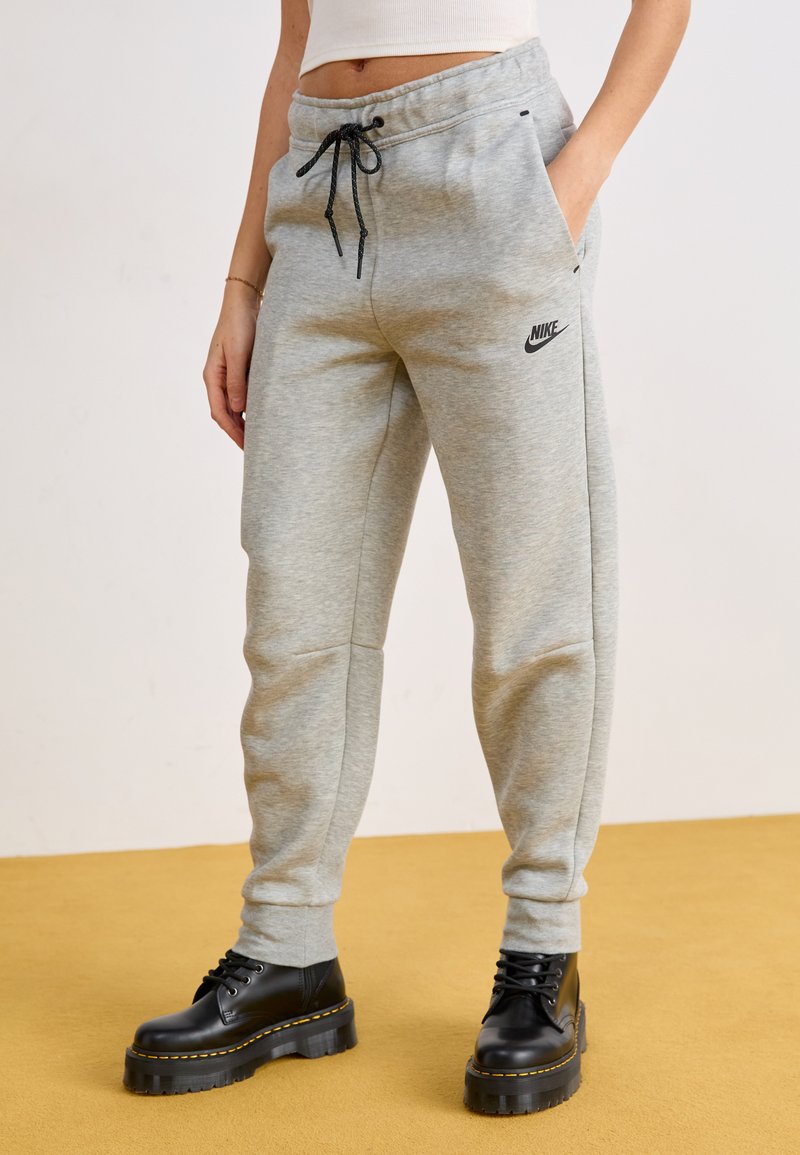 Pantalons de jogging en coton gris avec taille à cordon de serrage et poches latérales, arborant un logo Nike noir sur la cuisse gauche. Associés à des bottes à plateforme noires.