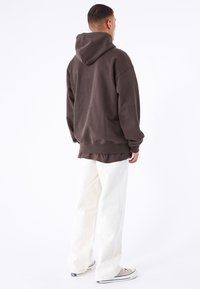 Brun oversized hoodie med huva och ribbade ärmslut, tillsammans med vita raka byxor; figuren står med ryggen vänd mot betraktaren.