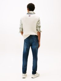 Suéter sin mangas de punto blanco sobre una camisa de cuadros, combinado con pantalones vaqueros azul oscuro y zapatillas grises. Presenta detalles de logo en el suéter.