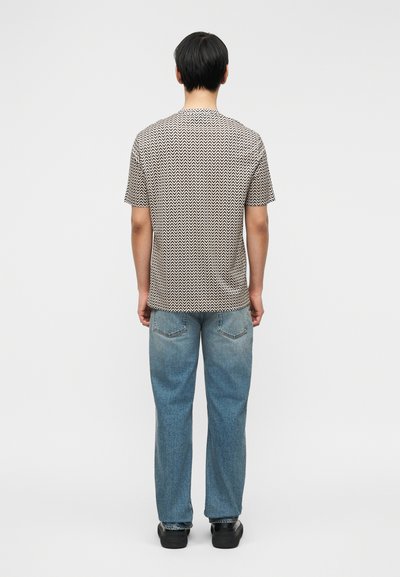 Άτομο με κοντά μαύρα μαλλιά που φοράει κοντομάνικο πουκάμισο με κρεμ και μαύρο μοτίβο, μπλε τζιν και μαύρα παπούτσια, όρθιο με την πλάτη προς τα εμπρός.