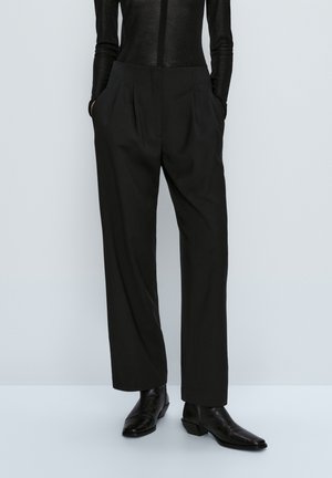 Personne portant un pantalon plissé noir taille haute avec des poches latérales, un haut noir cintré à manches longues, et des bottines noires à bouts pointus.