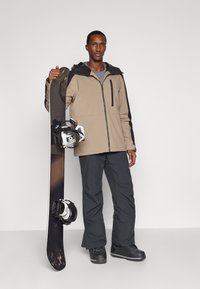 Άνδρας με μπεζ σακάκι και μαύρο παντελόνι κρατάει το σνόουμπορντ όρθιο μπροστά σε λευκό φόντο.