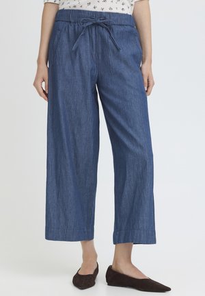 Vrouw die losse, cropped blauwe spijkerbroek draagt met een trekkoord in de taille, gecombineerd met donkere puntige platte schoenen op een lichte achtergrond.