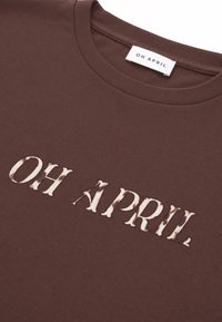 Κοντινό πλάνο καφέ T-shirt με στρογγυλή λαιμόκοψη και λευκό κείμενο "OH APRIL" τυπωμένο στο στήθος και ετικέτα της μάρκας στο εσωτερικό του γιακά.