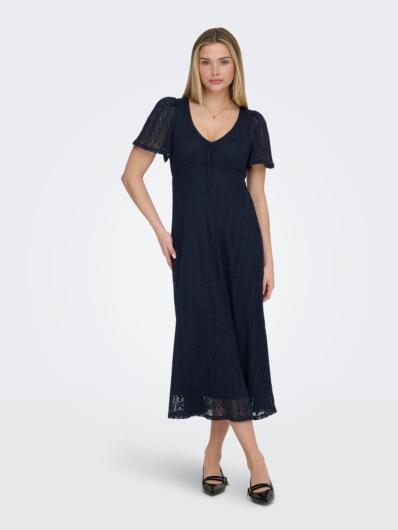 Žena stojaca v námornícky modrých midi šatách z čipky s krátkymi rukávmi a čiernymi lodičkami so špicatou špičkou, na jednoduchom svetlom pozadí.