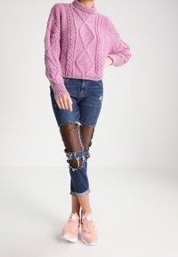 Růžový pletený svetr s texturovanými vzory, modré odrbané džíny s rozevřeným detailem na kolenou a světle růžové sportovní boty s bílými akcenty.