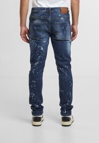 Mörkblå denimjeans med smal passform, med vita splattermönster, fem fickor och en brun läderlapp på midjan.