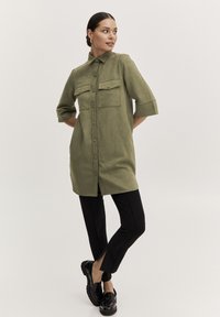 Abito camicia verde oliva, con bottoni e maniche corte, dotato di due tasche sul petto e una vestibilità morbida, abbinato a pantaloni neri slim e mocassini neri.
