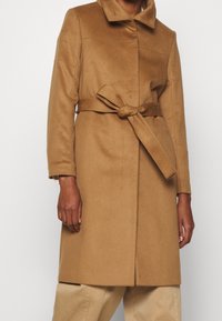 Person iført en mellemlang kamelcoat med et bundet bælte i taljen og beige bukser foran en ensfarvet baggrund.