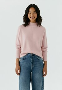 Jeune femme aux cheveux foncés mi-longs portant un pull rose clair et un jean en denim bleu, souriant devant un fond blanc uni.