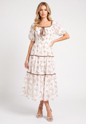 Femme portant une robe midi blanche avec un imprimé papillon marron, manches bouffantes, jupe à volants et encolure nouée, posant avec une main sur la hanche.